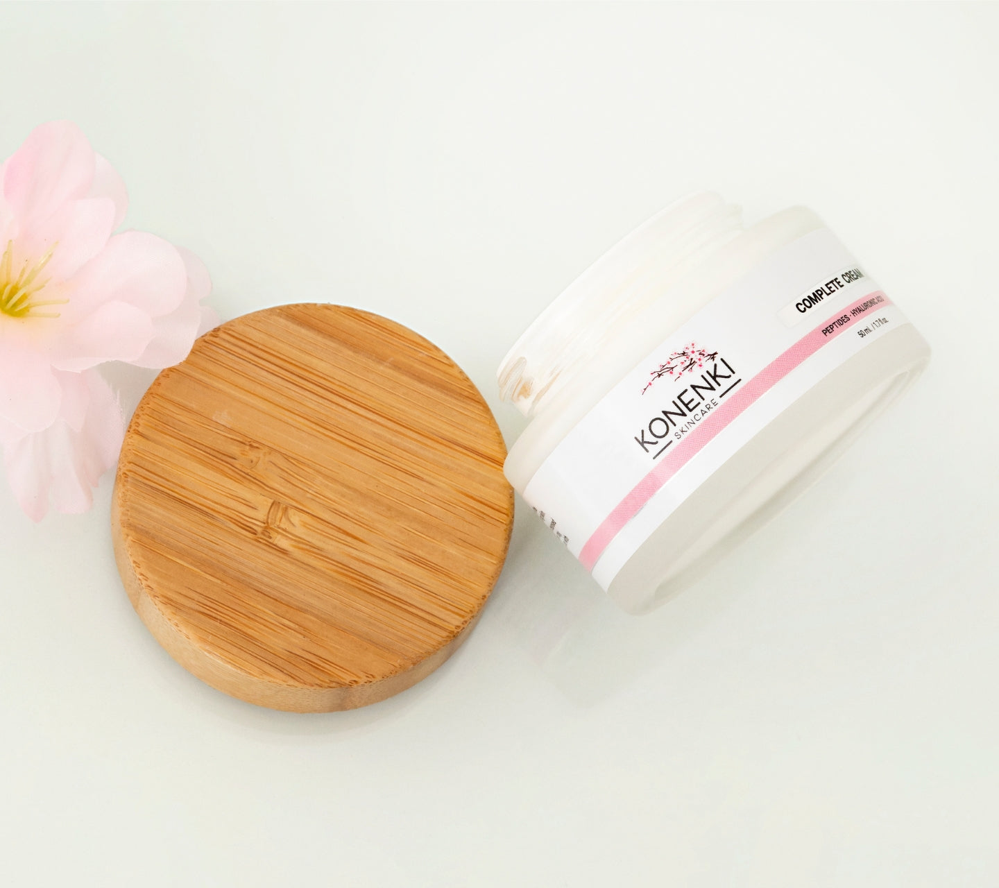 A bottle of Konenki Skincare's Complete Cream next to a pink flower against a blank white background.