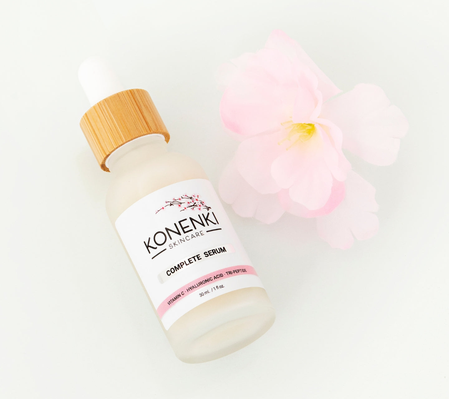A bottle of Konenki Skincare's Complete Serum next to a pink flower against a blank white background.