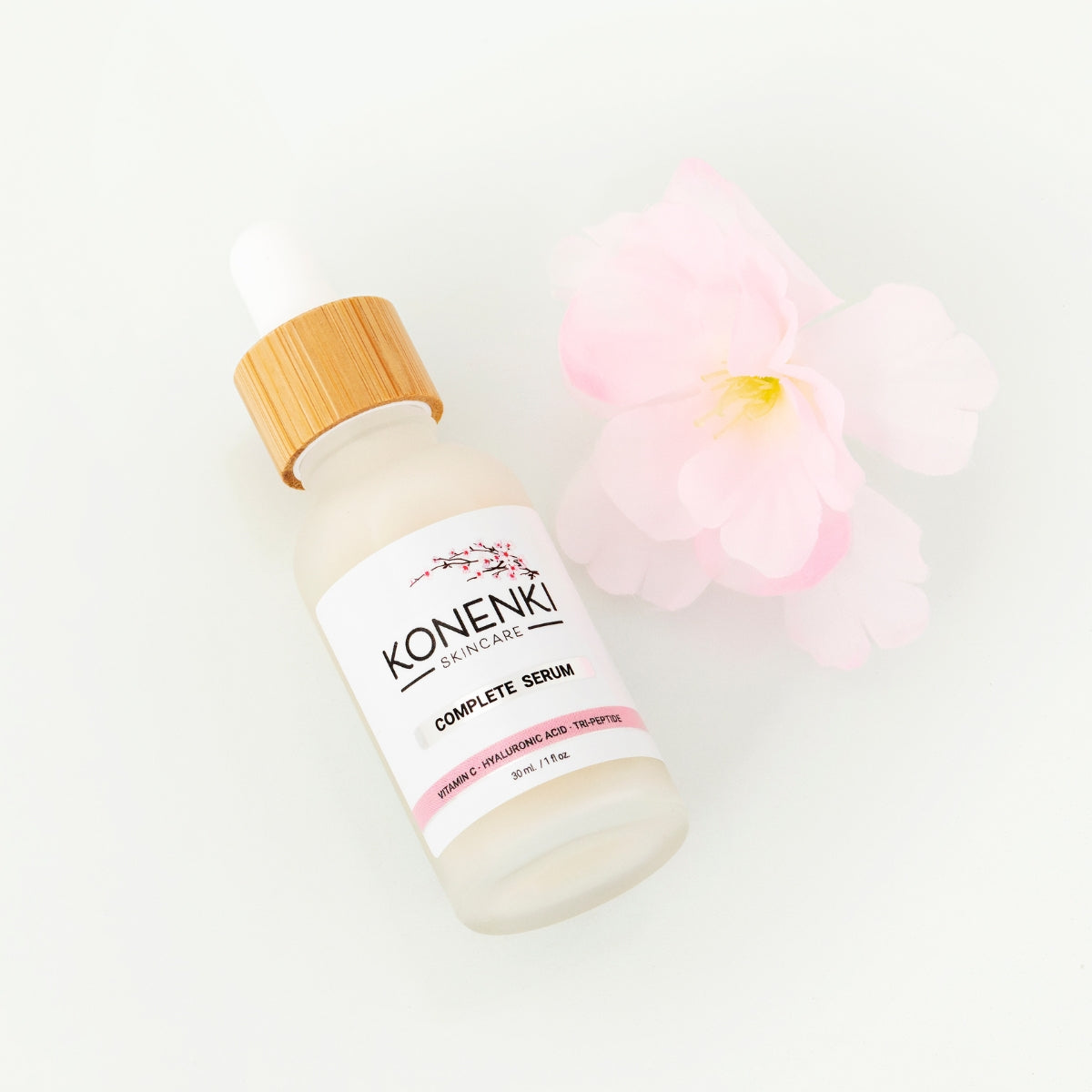 A bottle of Konenki Skincare's Complete Serum next to a pink flower against a blank white background.