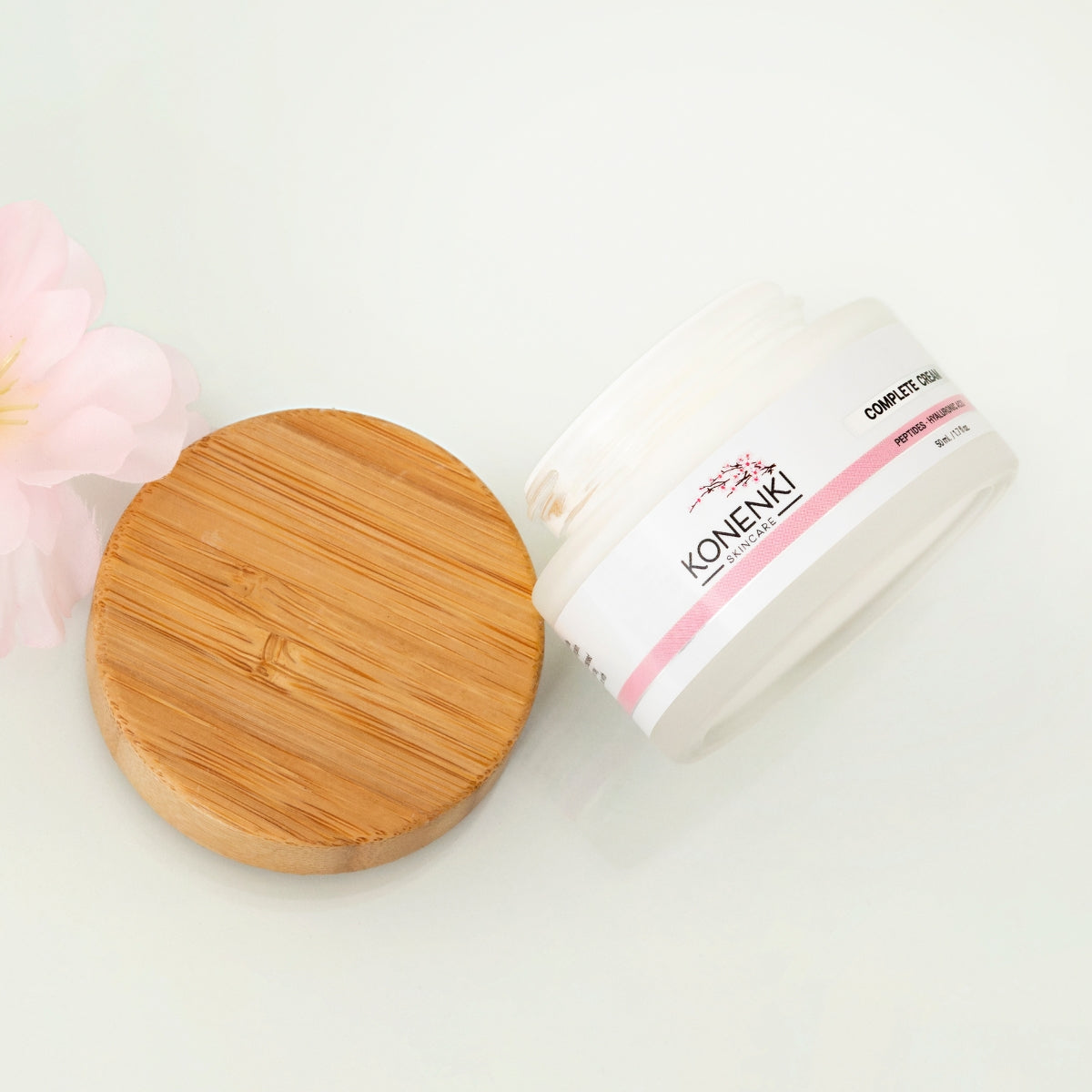 A bottle of Konenki Skincare's Complete Cream next to a pink flower against a blank white background.