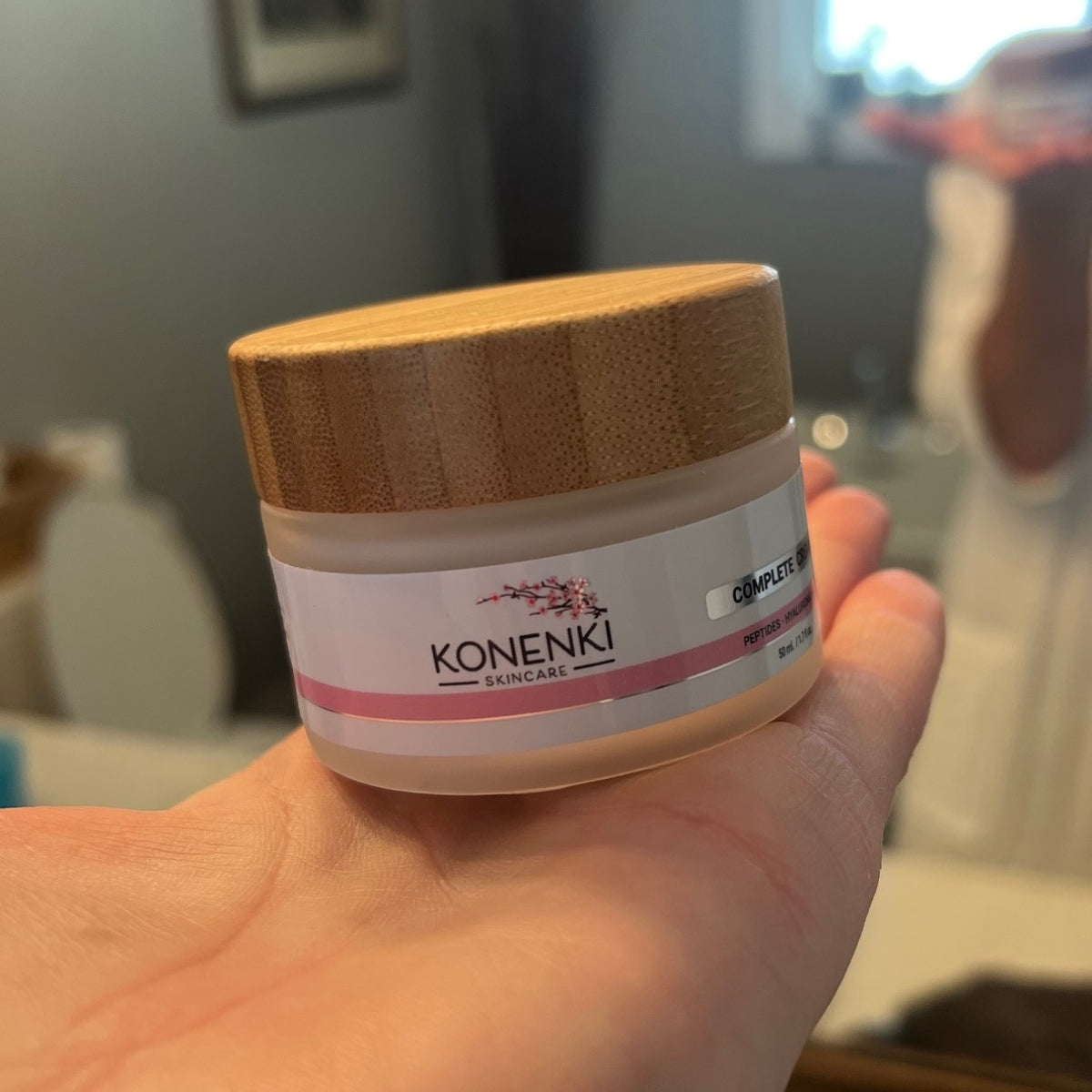 A woman's hand holds a bottle of Konenki Skincare's Complete Cream up to the camera, a bathroom mirror visible in the background.