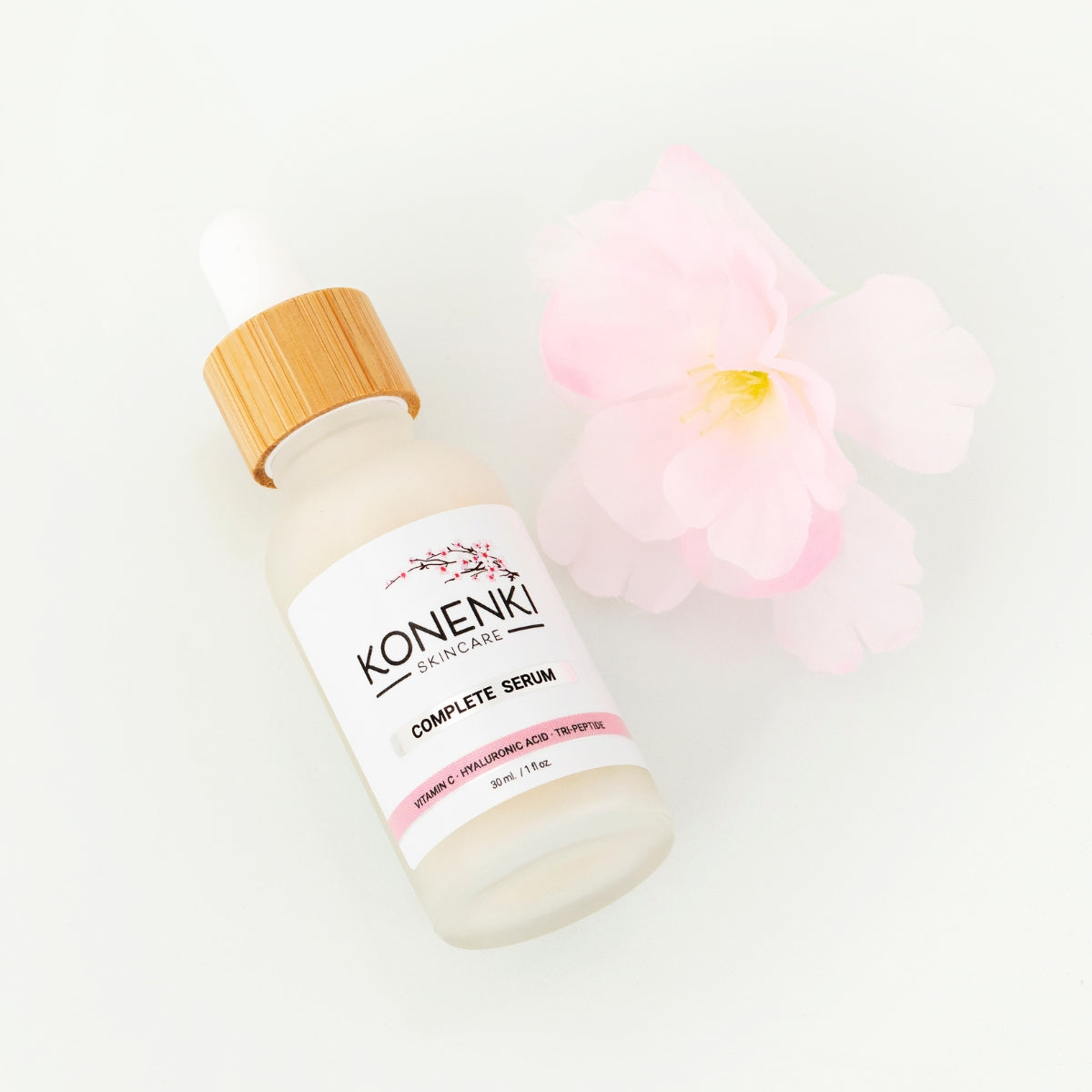 A bottle of Konenki Skincare's Complete Serum next to a pink flower against a blank white background.