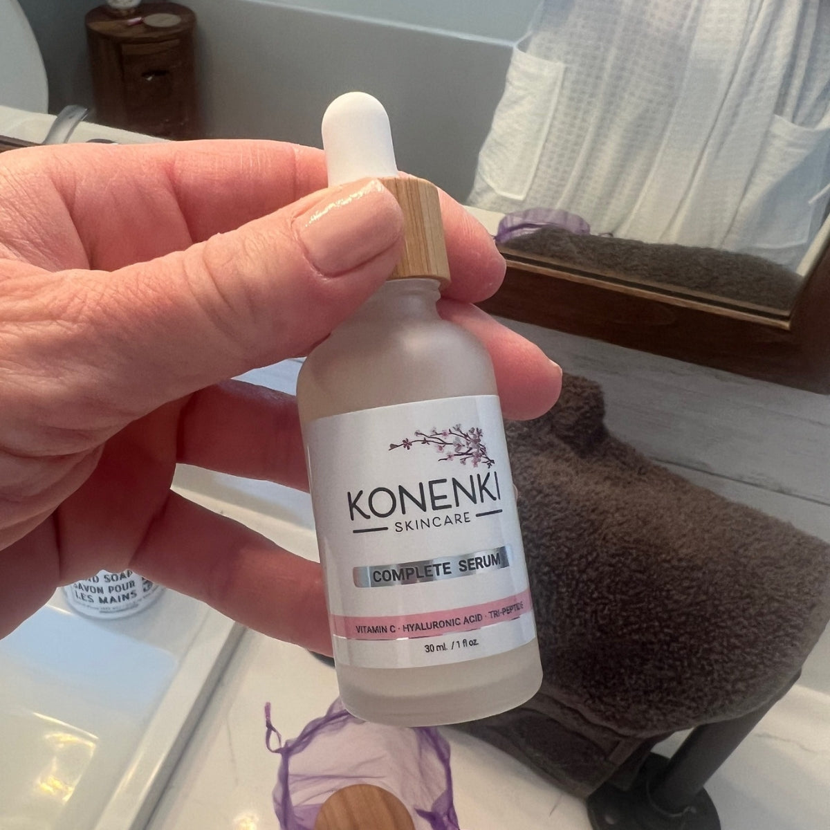A woman's hand holds a bottle of Konenki Skincare's Complete Serum up to the camera, a bathroom mirror visible in the background.