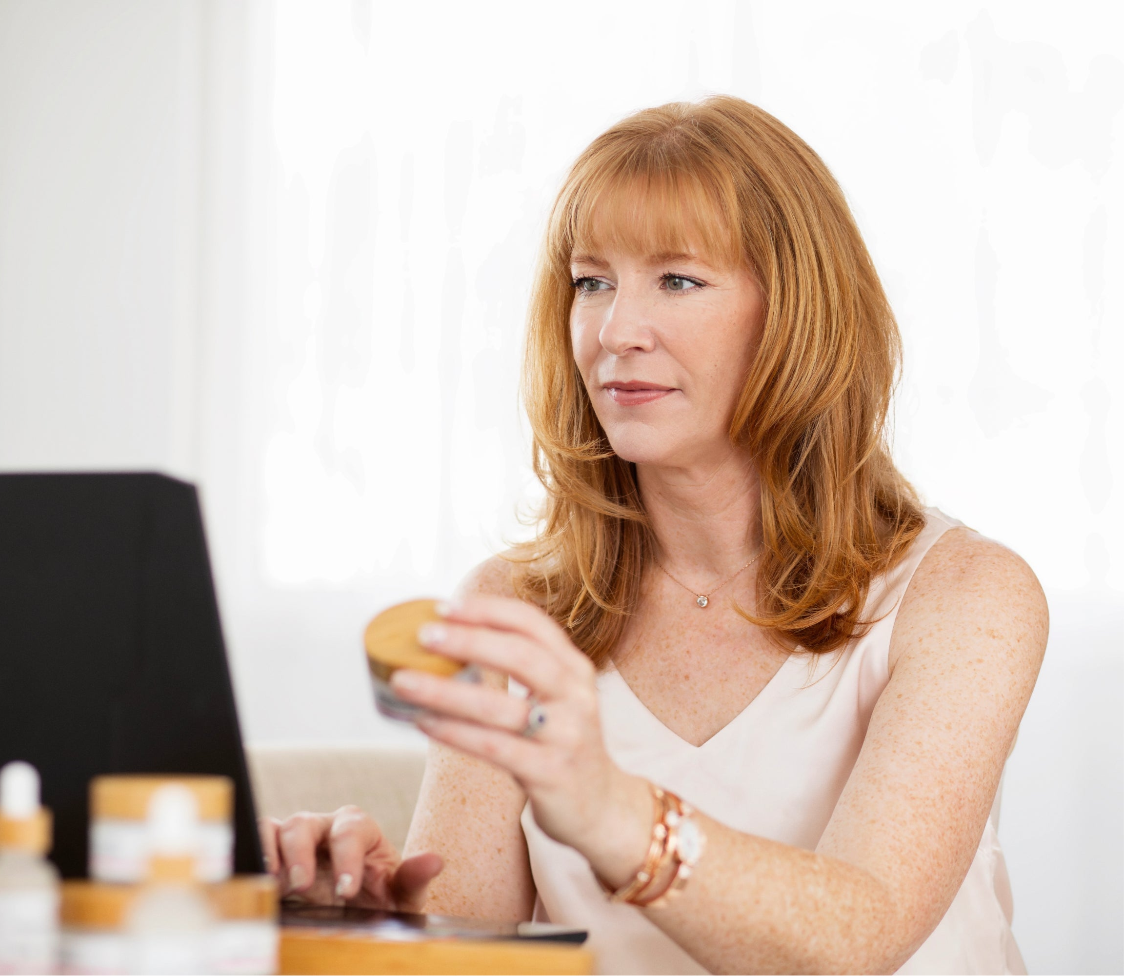 Konenki Skincare founder, Alana Palmer, sitting at a desk and looking at a laptop while holding a bottle of the Konenki Complete Cream.