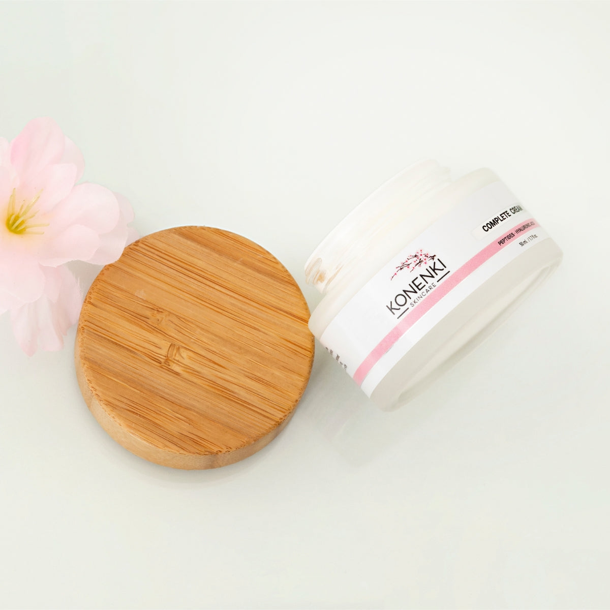 A bottle of Konenki Skincare's Complete Cream next to a pink flower against a blank white background.